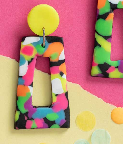 Nahaufnahme eines Ohrrings aus FIMO mit bunten Neon-Flecken auf Schwarz und gelbem rundem Stecker.