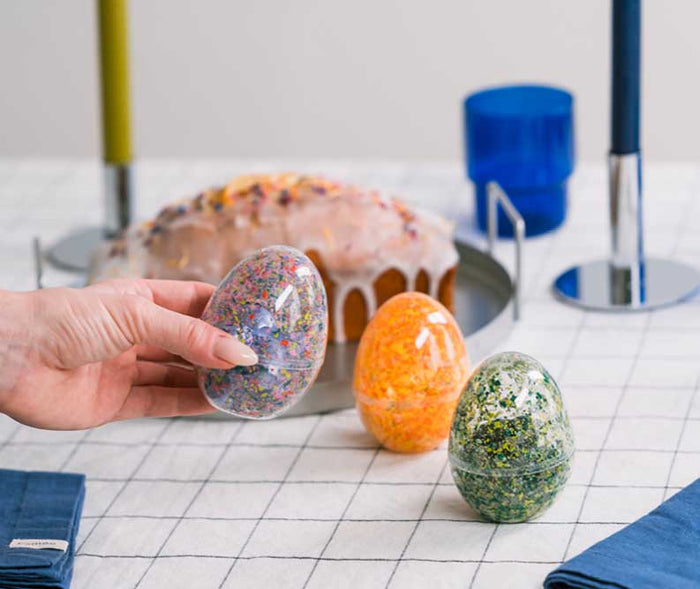 Hand präsentiert drei selbst gestaltete Überraschungseier mit bunten Farbmustern aus Wachsmalstiften. Die dekorativen Kunststoff-Eier stehen auf einem festlich gedeckten Ostertisch mit Kerzen und Osterkuchen.