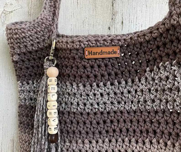 Selbst gehäkelte Tasche im Farbverlauf von Beige zu Braun hängt dekorativ an einer Holzwand und ist mit einem „Handmade“-Label sowie einem personalisierten Schlüsselanhänger verziert.