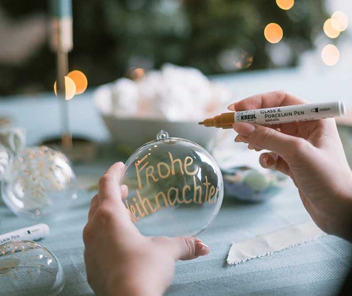 Eine transparente Weihnachtskugel wird mit einem goldenen Glas- und Porzellanmarker mit der Aufschrift „Frohe Weihnachten“ individuell beschriftet.