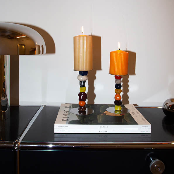 DIY Kerzenständer aus FIMO mit bunten Kugeln und brennenden Stumpenkerzen auf Zeitschriften auf einem schwarzen Sideboard.