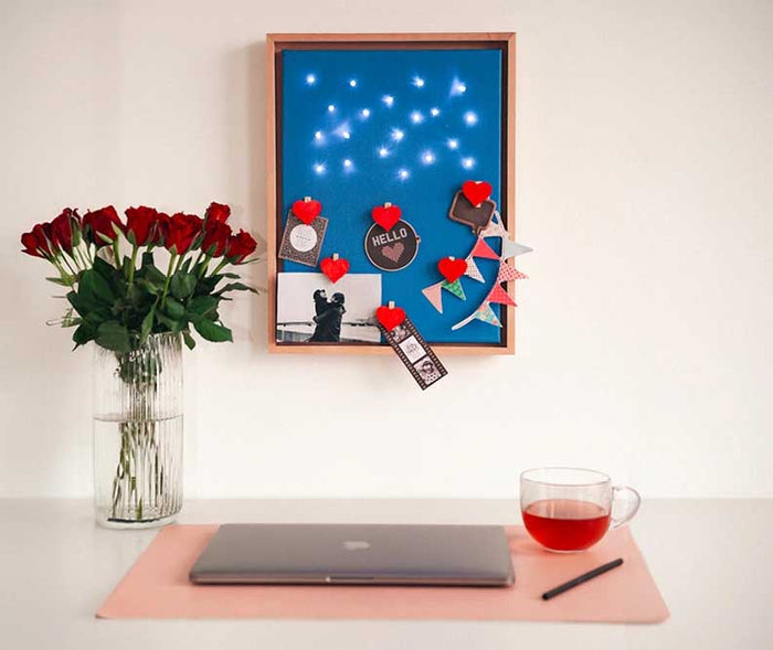 Modern eingerichteter Arbeitsplatz mit blauem Memoboard, dekoriert mit Fotos, roten Herzklammern, Lichterkette und Girlande. Daneben stehen rote Rosen, Laptop und eine Tasse Tee.