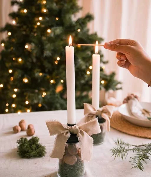 Hand zündet weisse Stabkerzen auf Gläsern mit Moos und Nüssen an, verziert mit beigen Stoffschleifen, vor einem beleuchteten Weihnachtsbaum