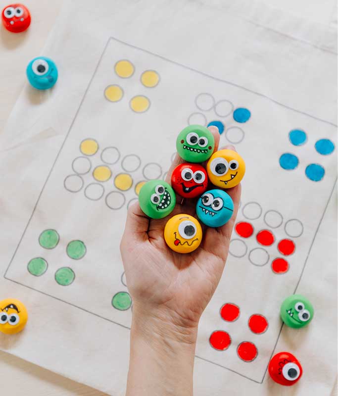 Hand hält sechs bunte Monster-Spielfiguren mit Wackelaugen über einem selbst gezeichneten Ludo-Spielfeld auf einer Stofftasche.