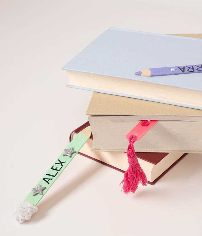Stapel Bücher mit personalisierten Lesezeichen aus Holzstäbchen. Die bunten DIY-Lesezeichen sind mit Namen, Quasten, Pompons und Glitzersternen verziert.