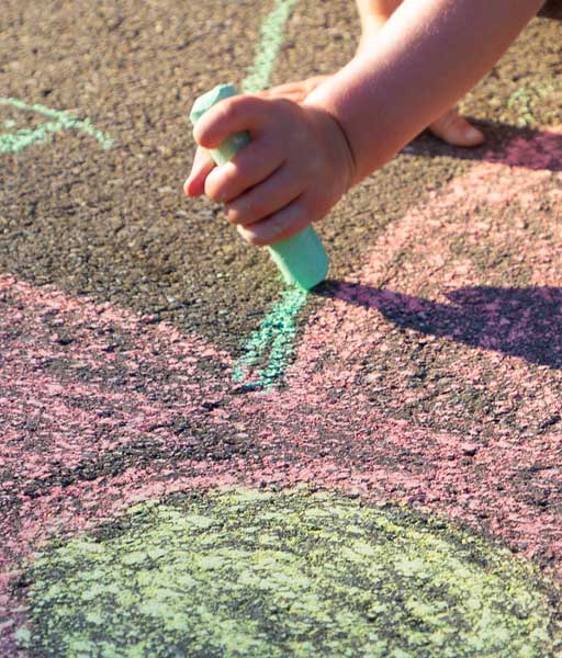 Kinderhand malt mit grüner Strassenkreide eine Linie auf den Asphalt, umgeben von rosa und gelben Kreidezeichnungen