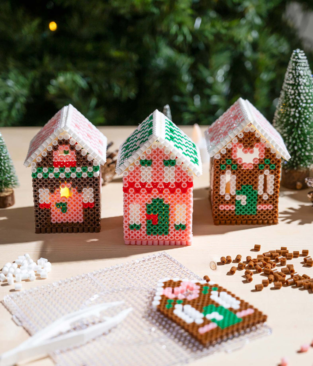 Drei weihnachtliche Lebkuchenhäuser aus bunten Bügelperlen mit detaillierten Mustern und Dächern auf einem Basteltisch, umgeben von Bügelperlen und Bastelzubehör.