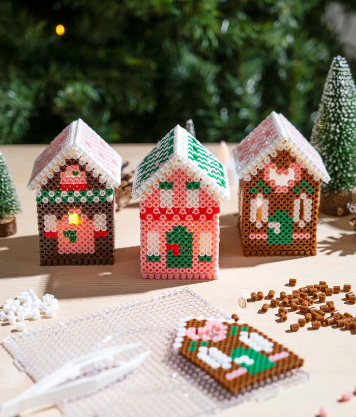 Drei weihnachtliche Lebkuchenhäuser aus bunten Bügelperlen mit detaillierten Mustern und Dächern auf einem Basteltisch, umgeben von Bügelperlen und Bastelzubehör.