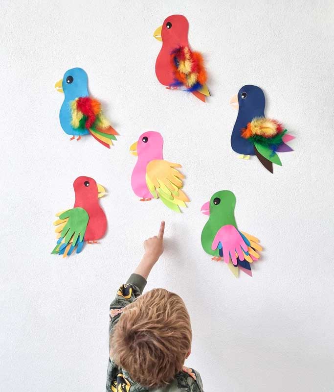 Ein Kind zeigt auf drei selbst gebastelte Papageien aus buntem Tonpapier, die an einer weissen Wand hängen. Die farbenfrohen Vögel sind mit handförmigen Flügeln und bunten Schwänzen gestaltet.