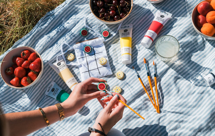 Sommerpicknick mit DIY Tic Tac Toe: Hände bemalen Wassermelonen-Spielsteine, daneben I AM CREATIVE Acrylfarben, Pinsel, Erdbeeren, Kirschen und Aprikosen