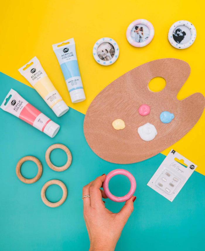 DIY-Bastelmaterialien mit Acrylfarben, Holzringen, Fotobuttons, Pinseln und Holzpalette für kreative Fotomagnete auf gelb-türkisem Hintergrund.