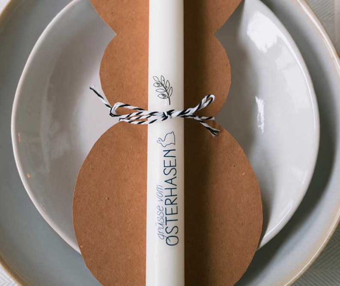 Weisse Osterkerze mit der Aufschrift „Grüsse vom Osterhasen“, befestigt auf einem Osterhasen-Anhänger aus Kraftpapier. Die Kerze ist mit einer schwarz-weissen Kordel dekoriert und dient als liebevolle Platzkarte oder Tischdekoration für Ostern.