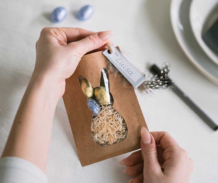 Eine Person hält eine selbst gebastelte Osterhasen-Geschenktüte aus Kraftpapier mit Hasenausschnitt. Die Tüte ist mit Holzwolle, Schokoladeneiern, einer Feder und einem personalisierten Namensschild gefüllt und eignet sich als liebevolle Osterverpackung o