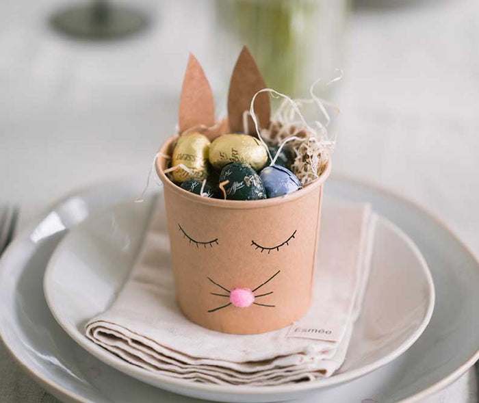Selbst gebastelter Osterhasen-Becher aus Kraftpapier, gefüllt mit Schokoladeneiern und Ostergras. Die niedliche Tischdekoration mit Hasenohren, geschlossenen Augen und rosa Pompon-Nase eignet sich perfekt als Osternest oder kleines Ostergeschenk.