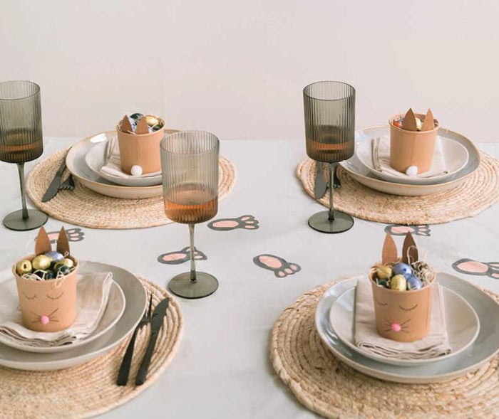 Festlich gedeckter Ostertisch mit Hasenbechern voller Schokoladeneier und Hasenpfoten-Spuren auf der Tischdecke. Kreative Osterdekoration für die ganze Familie.