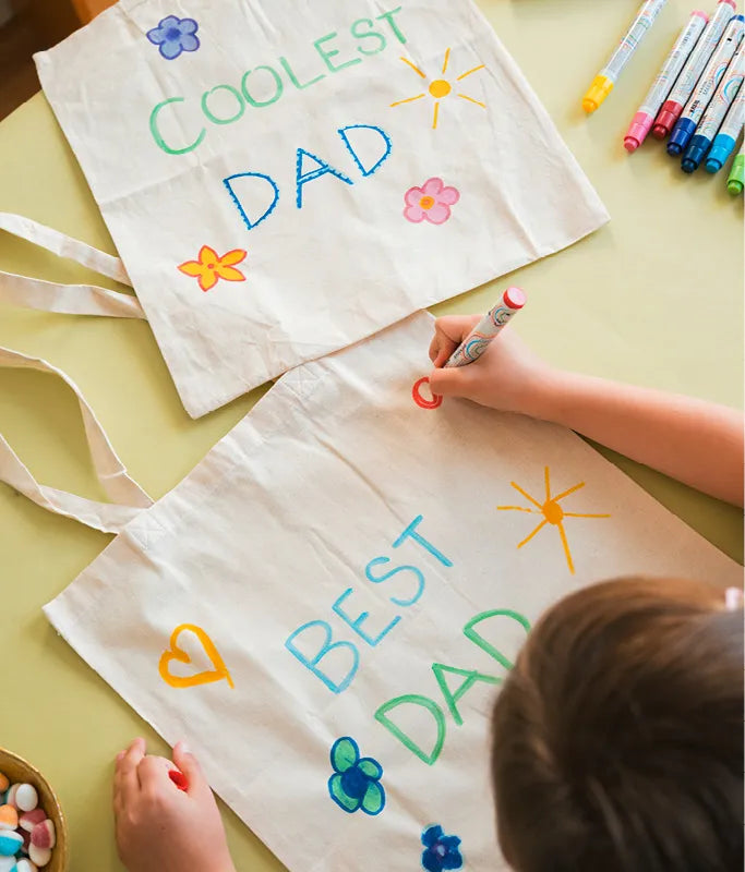 Kind bemalt eine Stofftasche mit bunten Motiven und dem Schriftzug „Best Dad“.