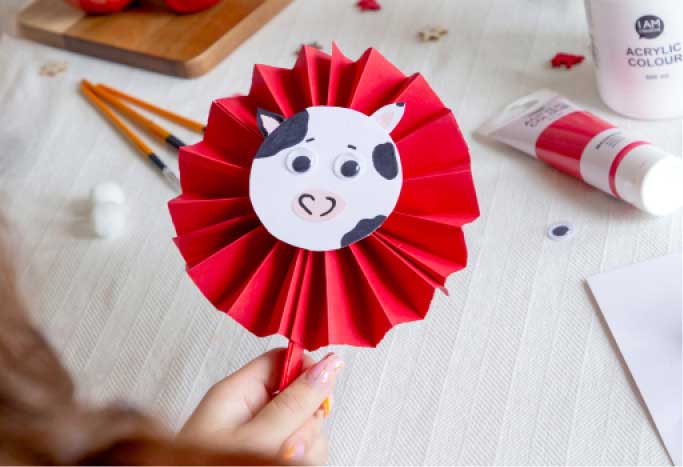 Hand hält einen selbstgebastelten Fächer aus rotem Faltpapier mit lustigem Kuhgesicht. Die kreative Schweiz-Dekoration eignet sich perfekt für den 1. August und sommerliche Festtage.
