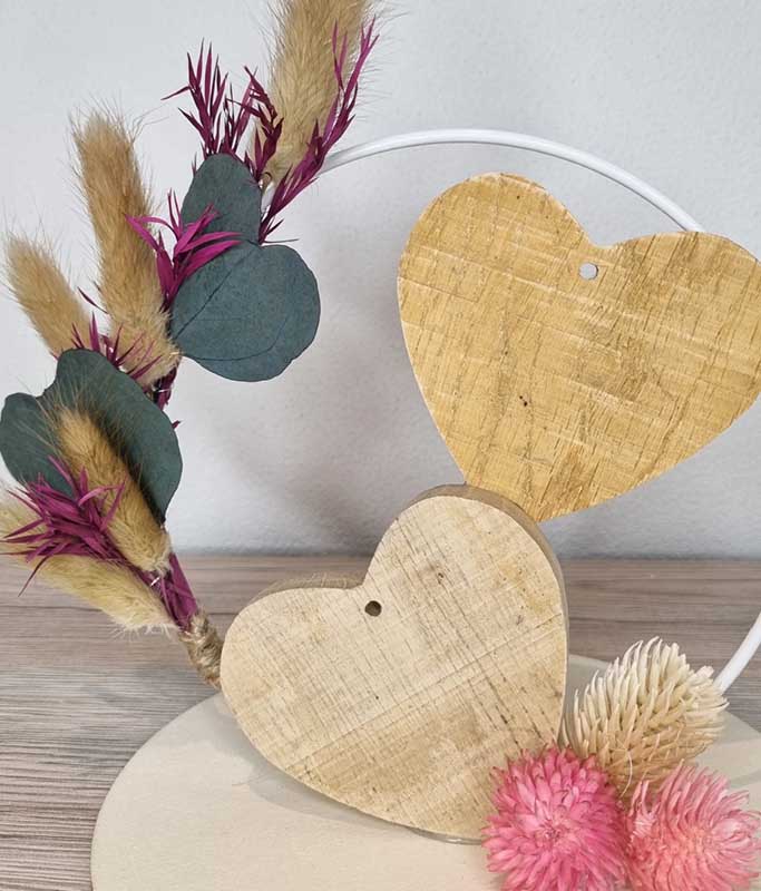 Dekorativer Herzring aus weissem Metall mit zwei Holzherzen, Trockenblumen, Eukalyptusblättern und rosa Trockenblüten. Die natürliche Kombination aus Holz und Trockenblumen macht die DIY-Dekoration zu einem stilvollen Blickfang.